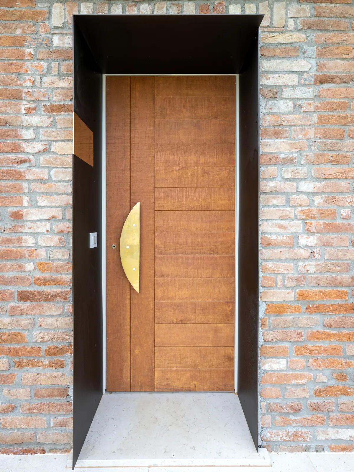 Portoncino in teak con maniglione a mezzaluna in ottone, incorniciato dall’imbotte metallico
